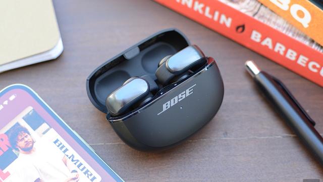 A pair of Bose Ultra Open Earbuds both sit in their charging case with the lid open on a wooden desktop surrounded by books, a smartphone and a pen.