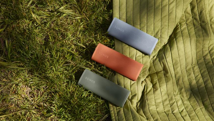 Three Sonos Roam 2 portable speakers (green, red, blue), sitting side-by-side (diagonal to the camera) on an outdoor blanket on top of grass.