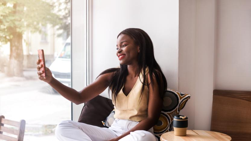 Attractive african girl makes selfie at the windowsill at cafe with reusable cup of coffee.