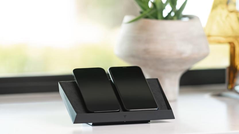 The Tesla Wireless Charging Platform with two phones on it, sitting on a white marble-like surface with a window and potted plant in the background.