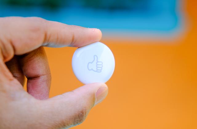 A hand holds a white circular object with a gray thumbs up on it. 