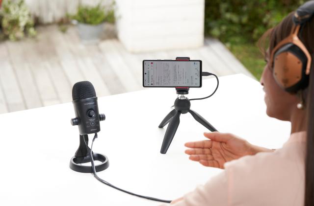 Lifestyle marketing photo of a person (view from behind, only part of them is visible in the right of the frame) talking into a Rode Go:Podcast microphone with a smartphone set up on a mini tripod next to it. White table with a blurred environment behind it.