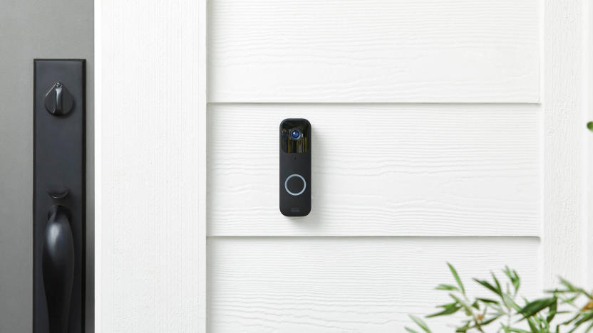 Marketing photo of a Blink Video Doorbell (horizontally oblong device, black, with a camera sensor on top and blue-ring button on bottom) on a white panel outdoor wall next to a front door.