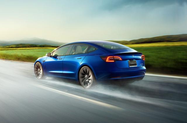 A blue Tesla Model 3 seen driving on a highway with a water-soaked surface blurred with speed.