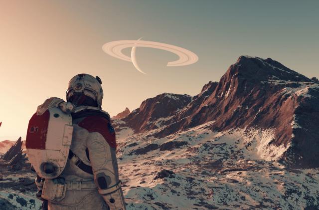 Media image (screenshot) from the game "Starfield." A person in an astronaut suit stands on a planet with reddish-brown mountains. A ringed Saturn-like planet is visible in the daytime sky.