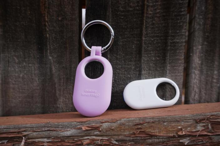 Two Samsung Galaxy SmartTag 2 Bluetooth trackers sit against a wood backdrop, one pink and the other white.