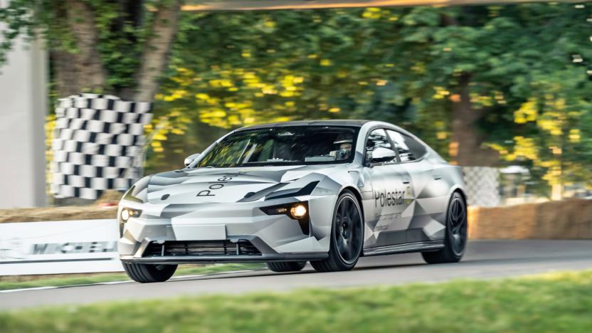 The upcoming Polestar 5 EV in grey with geometric patterns is seen on a racetrack, with grass in the foreground and trees behind it with checkered tape around them.