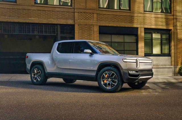 A silver Rivian R1T EV pickup truck parked out front of a brick building late in the day with shadows and low evening sun.