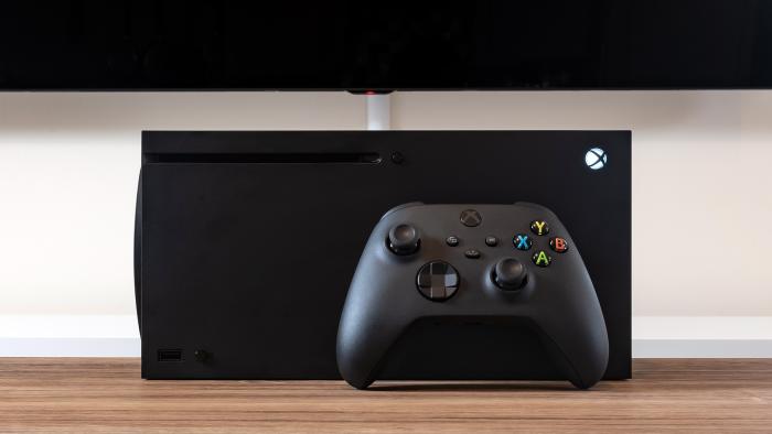 The Xbox Series X game console lays on its side, with a black Xbox controller rested in front of it, on top of a brown wooden desk.