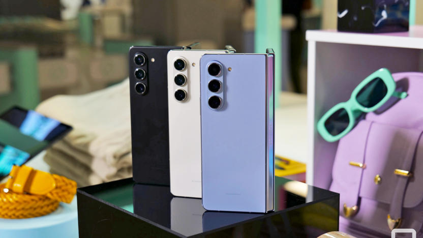 Three of the Samsung Galaxy Z Fold 5 phones stand on end open 45-degrees and showing the exterior of the phone, while sitting amongst a variety of pastel colored props, bags, sunglasses and folded clothing.