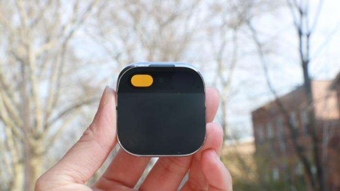 The Humane AI Pin held in mid-air in front of some bare trees and a street with red brick buildings on it.