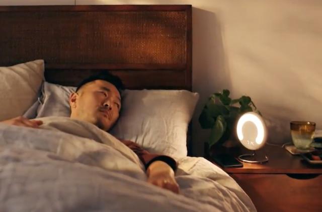 A man in bed with a round lamp on the nightstand next to him.