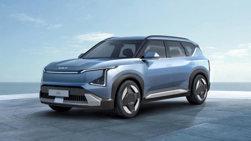 A light blue Kia EV5 SUV is parked on a floor of large tile slabs with the ocean and a blue sky in the background.