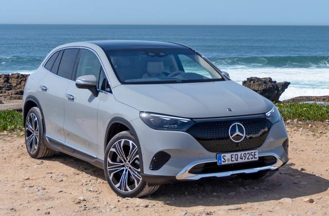 A grey Mercedes EQE SUV is parked on a rocky shore with rock outcroppings and waves close at hand in the background.