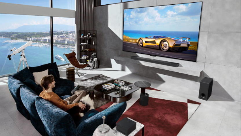 A man sits on a blue couch with his dog watching a race car on his humongous LG TV. It's a modern bachelor pad overlooking a bay with a telescope by the large windows.