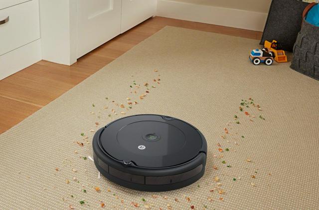 A robot vacuum is picking up small pieces of colorful debris on a tan run in a child's room. There's a small bulldozer toy and a toy basket in the background. 