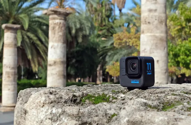 The GoPro Hero 11 Black Mini pictured on a rocky surface in a park with roman-style columns and palm trees in the background.