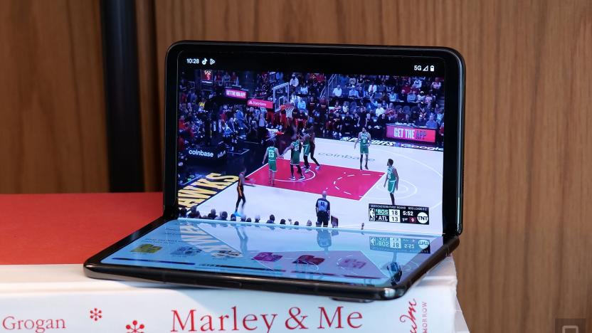The Google Pixel Fold in "tabletop view" half opened atop a stack of books. A basketball game is displayed on the upper screen. Apps and other icons are visble on the bottom screen. 