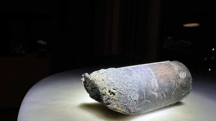 Photo of a stanchion from an ISS space dump. The object, a gray-ish cylinder with crumbled / burned edges, sits on a white table with black background.