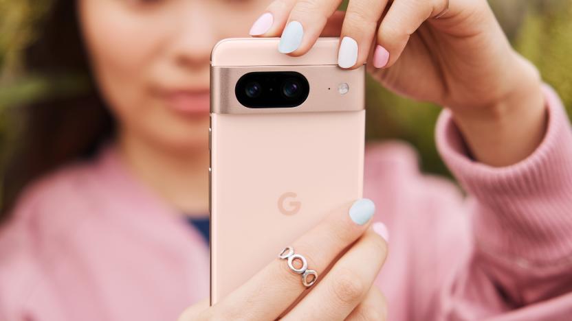 A woman in a pink sweater holds up a pink smartphone, the Google Pixel 8, in front of the camera, showing its back and camera system.