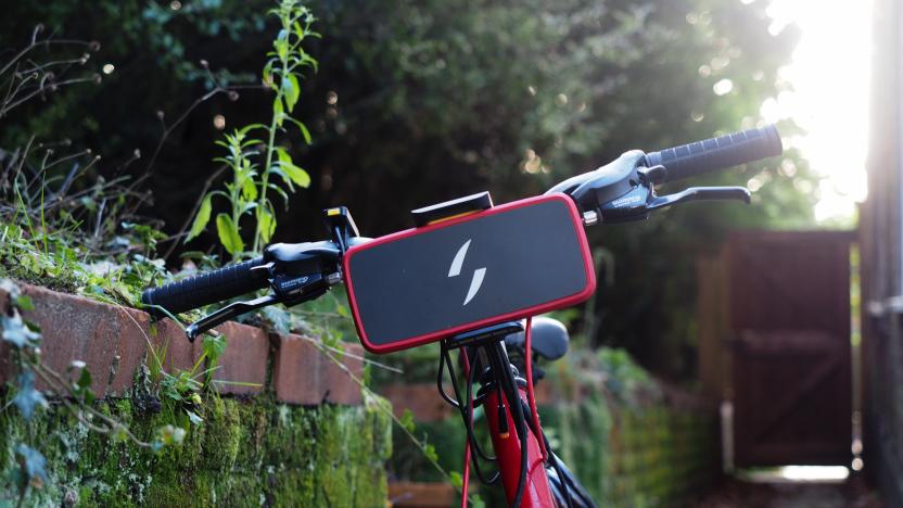 The front view of a red bike with the Swytch Air e-bike conversion kit installed, showing the battery pack portion attached to the front of the handlebars. The bike is leaning against a vine-covered wall.