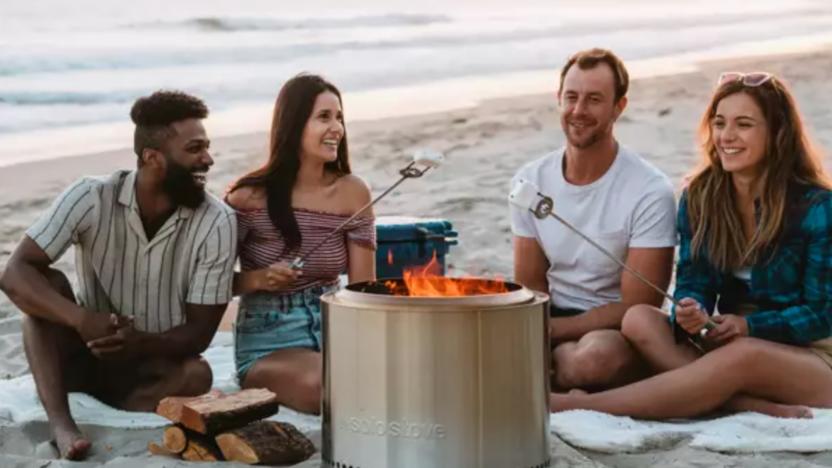 Some young hotties sitting around a fire pit laughing and roasting marshmallows. 