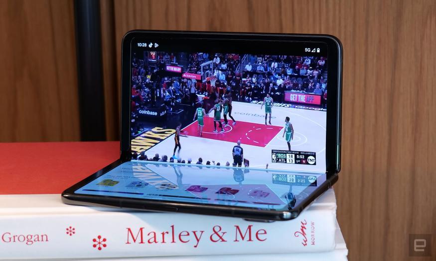 The Google Pixel Fold in "tabletop view" half opened atop a stack of books. A basketball game is displayed on the upper screen. Apps and other icons are visble on the bottom screen. 