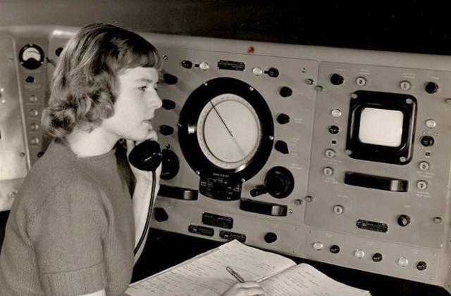 Archival black & white photo of Virginia Norwood, sitting in a (mid-20th-century) control room while talking on the phone and jotting notes in a log book.