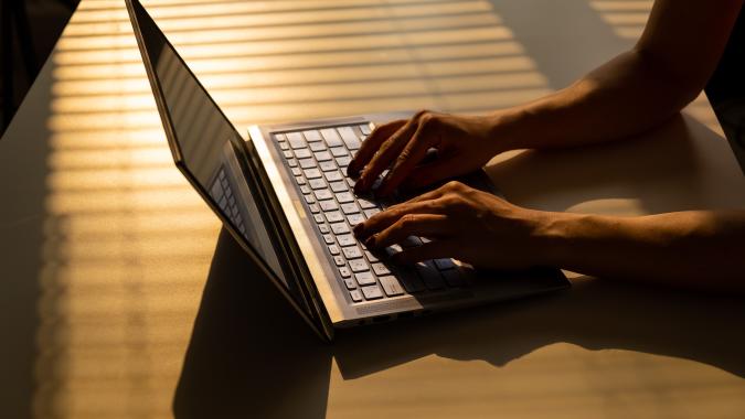 A woman is typing on a laptop keyboard on a white table. The shadow from the blinds falls on the desktop