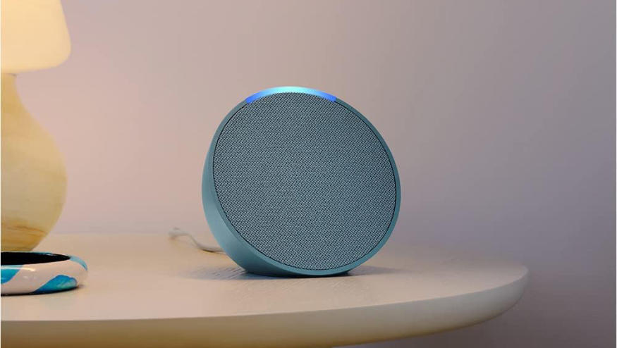 The Amazon Echo Pop speaker in blue sits on a night table with a yellow lamp nearby. 