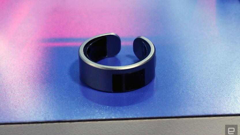 Image of the VTouch Whsp Ring, in dark grey, sat on a blue to pink ombre table.