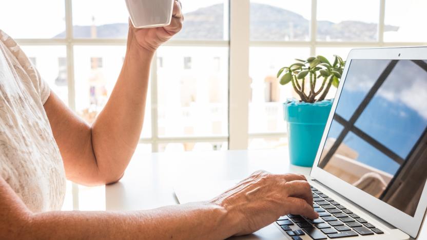 Close-up view of woman's hand typing on laptop keyboard while holding a cup of tea or coffee. Nomadic mature female enjoying tech and social working home