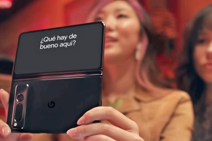 Still from a Google promotional video, featuring two women in a restaurant booth holding up a Pixel Fold phone (the phone is in focus, the women are blurred). The phone's outer screen shows the translation of her spoken words: "¿Qué hay de bueno aqui?"