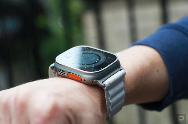 Off-angle view of the Apple Watch Ultra with alpine loop on a person's wrist. From this perspective, the orange Action button and speaker grill on the left edge are in view, while the screen's contents are dim.