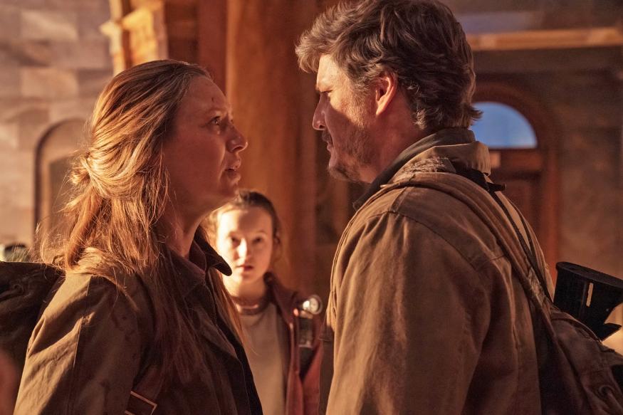 Tess (Anna Torv) and Joel (Pedro Pascal) stand face-to-face with serious expressions in The Last of Us. Ellie (Bella Ramsay) is slightly out of focus in the background.