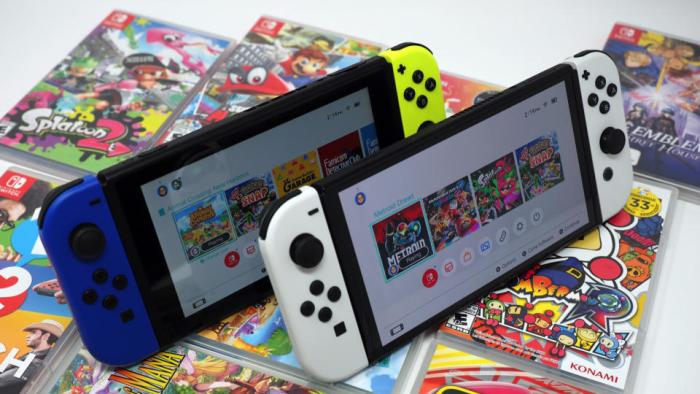 Two Nintendo Switch consoles sitting upright on an array of game boxes. Standard model with blue and yellow Joy-Cons on the left, OLED model with white Joy-Cons on the right.