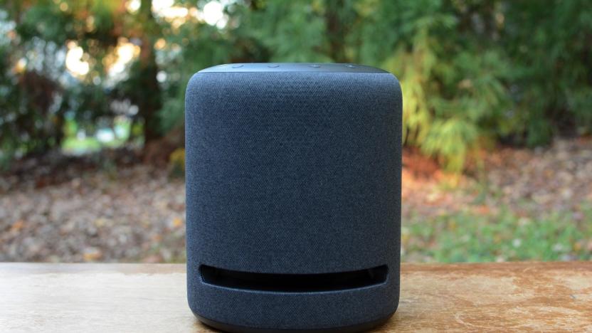 Amazon's Echo Studio Speaker on a wooden table outdoors in front of a bank of trees. 