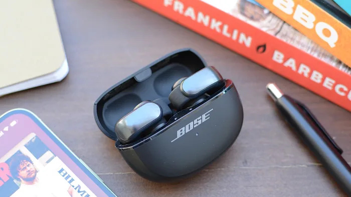 A pair of Bose Ultra Open Earbuds both sit in their charging case with the lid open on a wooden desktop surrounded by books, a smartphone and a pen.