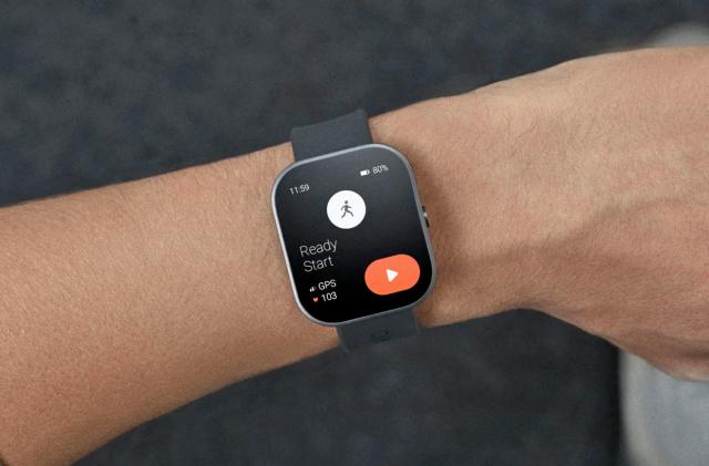 A rectangular-faced smartwatch with rounded edges and a grey band is shown in close-up on a person's wrist. 