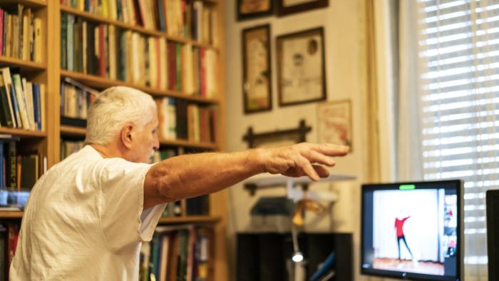 Side view of Senior man, a Parkinson Disease patient, who is standing  in front of monitor, at home, online exercising dancing with help of a physical therapist for Parkinson Disease. Online is only possible therapy as it is Covid-19 lockdown time.