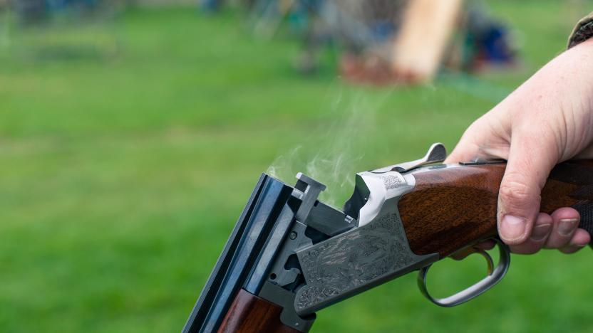 A close up of a person holding a shotgun with smoke coming out of the barrel with grass in the background.