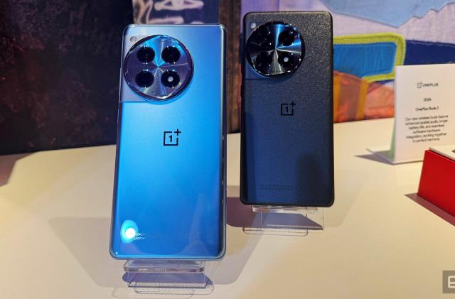 Two OnePlus 12 phones site on display stands at an event. Both have their rear camera facing forward, one is a light blue metallic and the other is a dark grey.