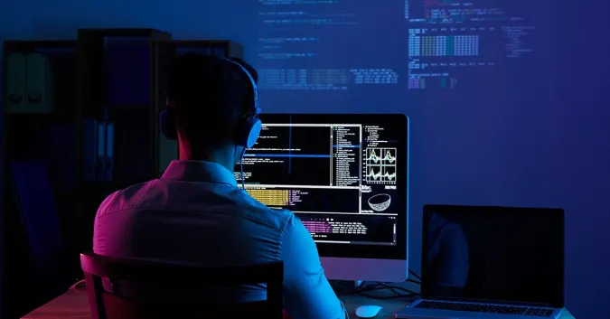 Stock image of someone using a computer in the dark with code projected onto the far wall.