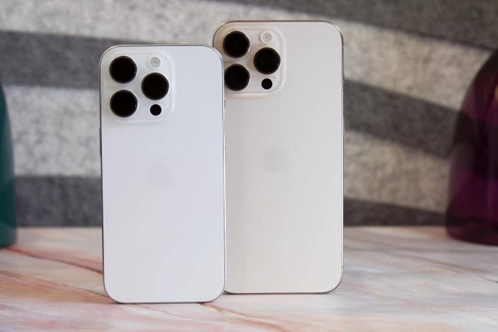 A white iPhone 16 Pro and a desert iPhone 16 Pro Max standing on a table.