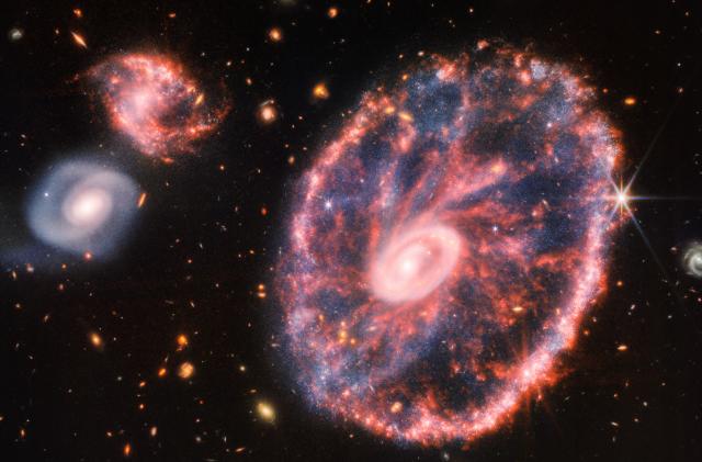 A large pink, speckled galaxy resembling a wheel with with a small, inner oval, with dusty blue in between on the right, with two smaller spiral galaxies about the same size to the left against a black background.