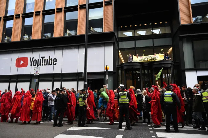 Climate activists hold a banner after climbing atop the roof of the entrance of the building and others line the pavement at ground level as they protest outside the UK office of Youtube during the tenth day of demonstrations by the climate change action group Extinction Rebellion, in London, on October 16, 2019. - Activists from the environmental campaign group Extinction Rebellion vowed Wednesday to challenge a blanket protest ban imposed by the London police. (Photo by Paul ELLIS / AFP) (Photo by PAUL ELLIS/AFP via Getty Images)