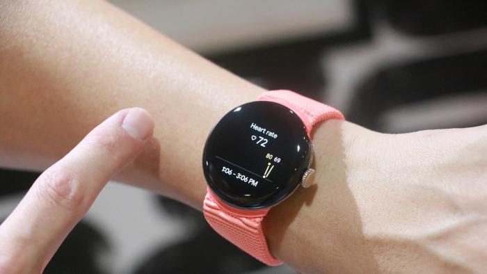 The Pixel Watch 2 with a peach woven band on a wrist, showing a heart rate reading.