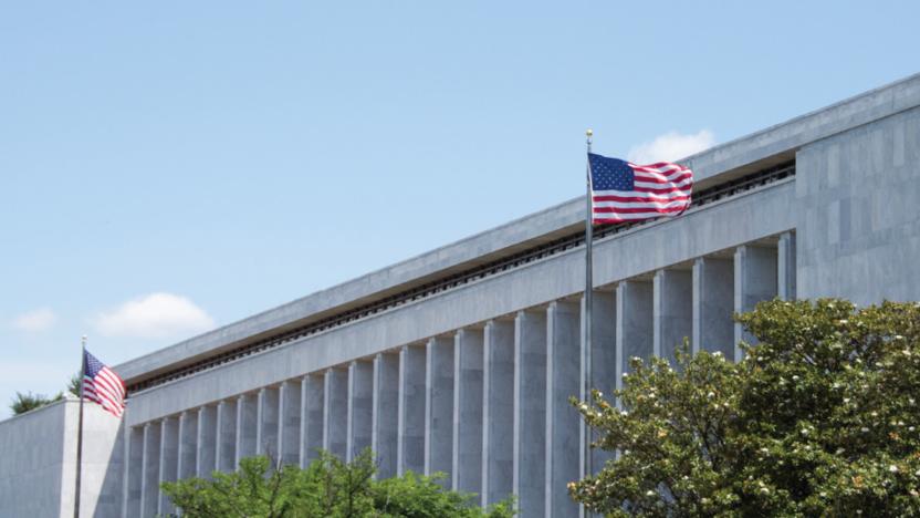 Outside view of a regal Washington, DC government building with a tall American flag and tree tops in front.