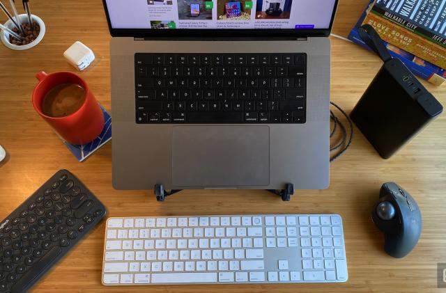 A MacBook Pro sits on a stand on a desk, surrounded by various keyboards and other useful accessories. 