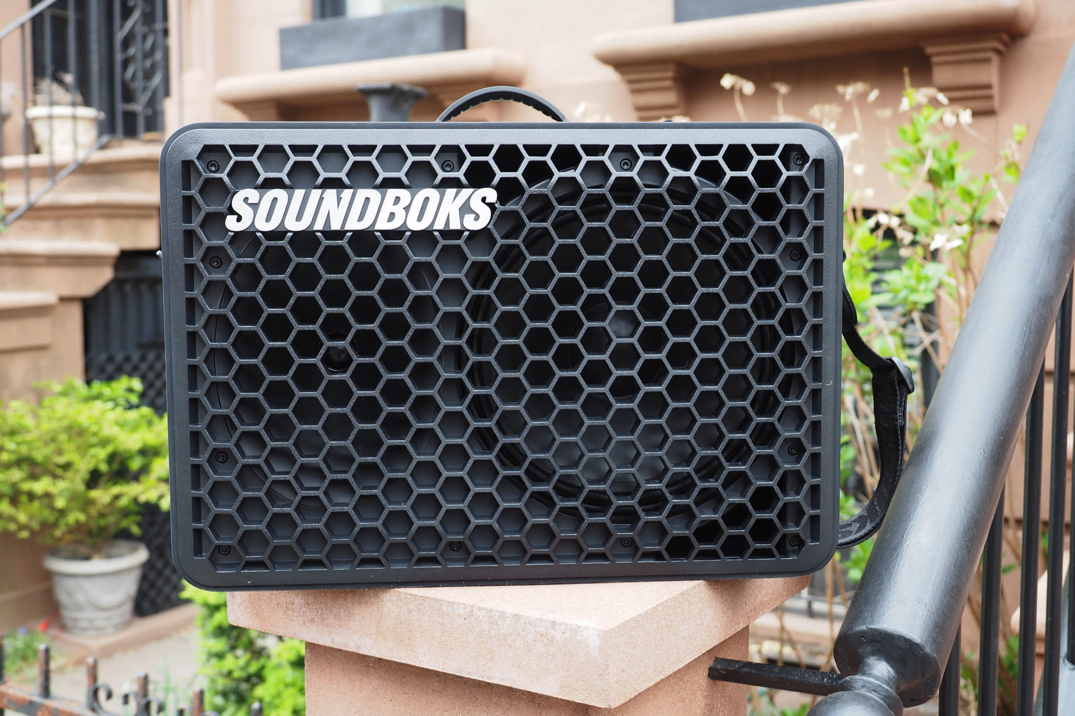The Soundboks Go portable Bluetooth speaker sitting in front of a Brooklyn brownstone, with plants, stairs and railings in the background.